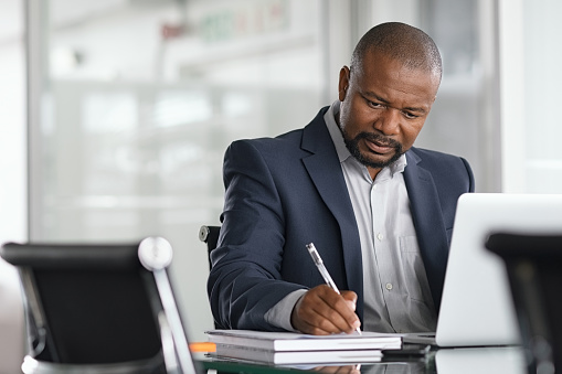 A focused man writes in a notebook with a laptop in front of him