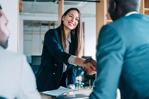 A woman shakes hands with a man across a table. She is holding a clipboard and smiling. 